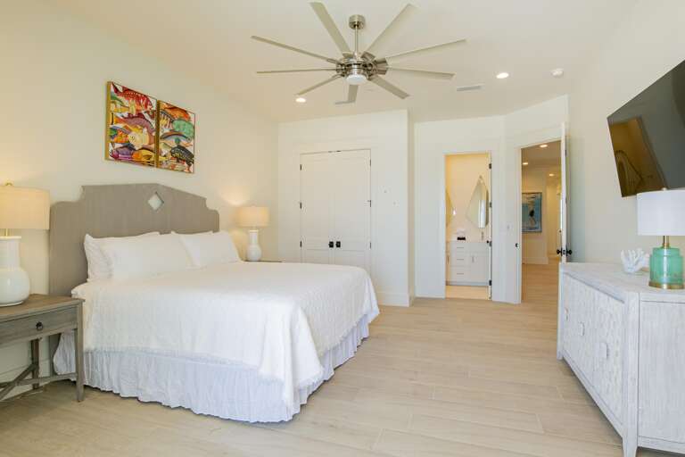 King bed with gray headboard between white lamps on wooden nightstands, light wood flooring, and wall-mounted TV.
