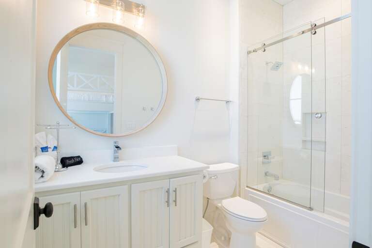 Bathroom with white beadboard vanity, round wood-framed mirror, and glass-enclosed tub with tiled surround.