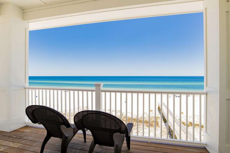 Covered balcony with dark woven chairs overlooks white sandy beach and turquoise Gulf waters.