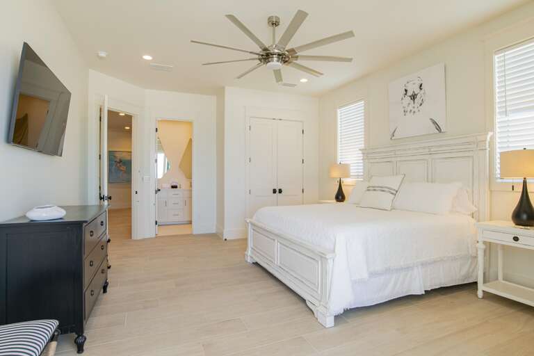 Bedroom with white distressed wood bed, two black lamps on side tables, light wood tile flooring, and ceiling fan.