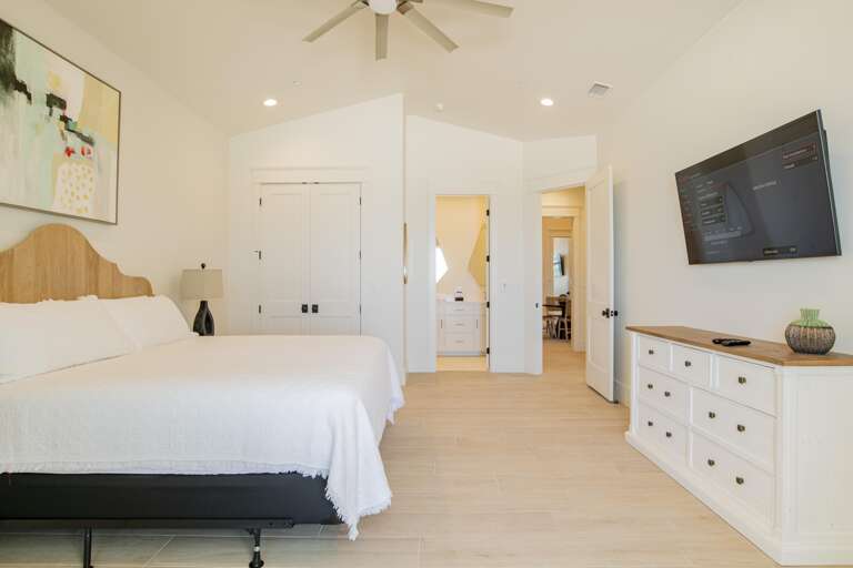 King bed with wooden headboard and white bedding sits opposite a wall-mounted TV above a white dresser topped with decor.