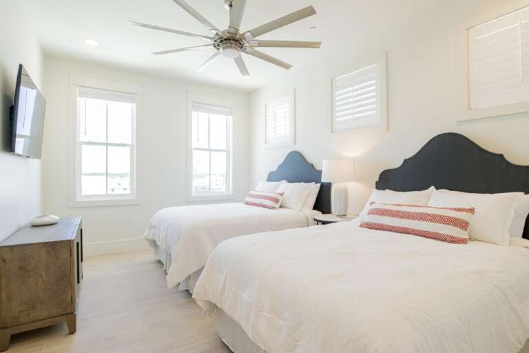 Two queen beds with navy headboards flank a bedside lamp, natural wood dresser, and wall-mounted TV.