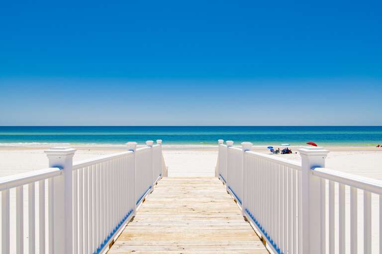 Boardwalk with white railings leads over light wood planks to a white sand beach and calm turquoise Gulf waters.