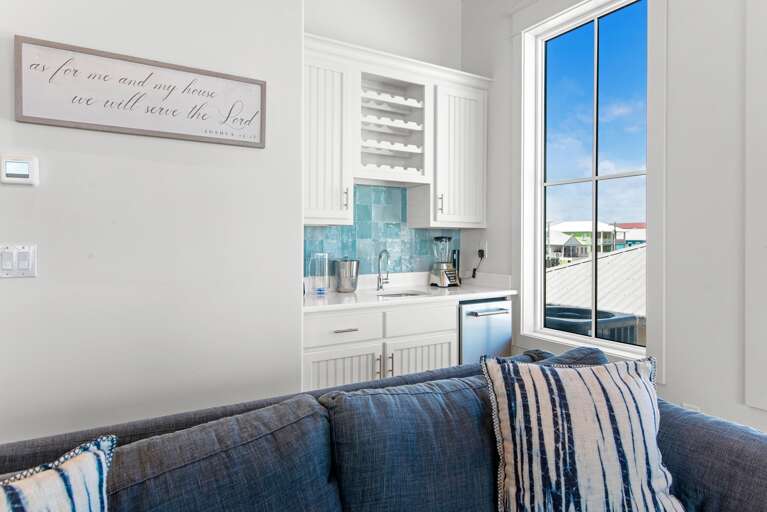 Wet bar with white cabinetry, turquoise tile backsplash, mini fridge, and large window beside a blue-gray couch.