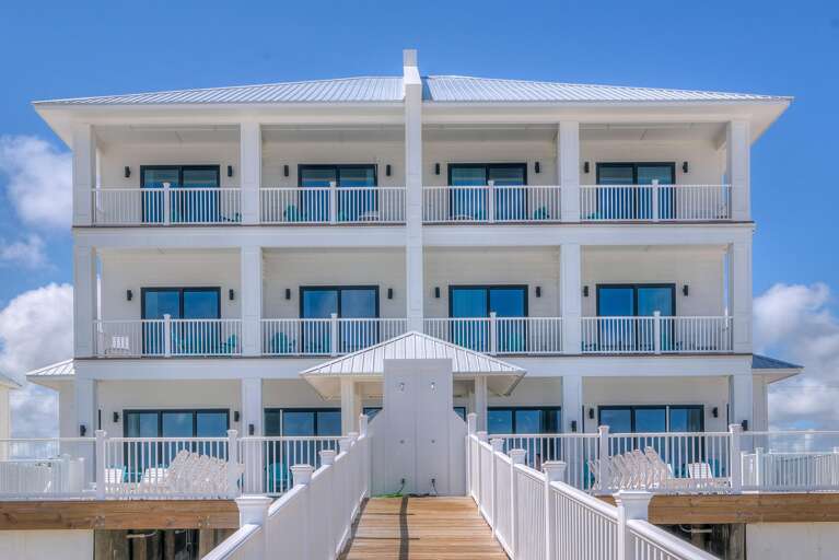 White three-story beachfront house with metal roof, wraparound balconies, and wooden ramp leading to entrance.