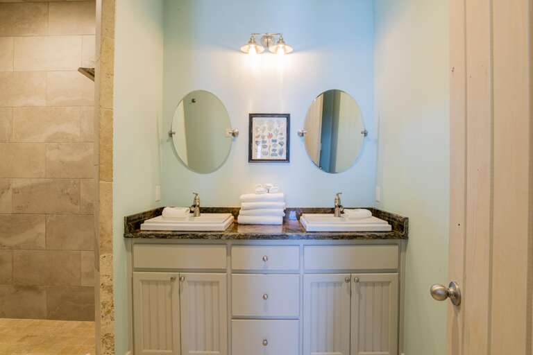 Dual rectangular sinks with brushed metal faucets sit on a dark granite countertop beneath oval mirrors and soft wall lighting.