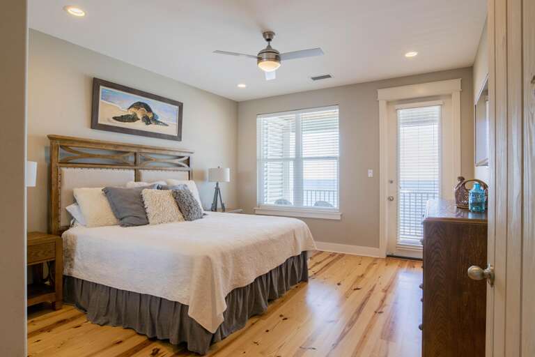 King bed with wooden headboard and textured pillows sits on polished pine floors beside window and balcony door.