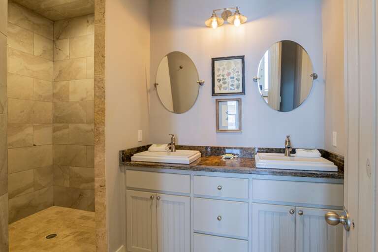 Bathroom vanity with dual white sinks, granite countertop, round mirrors, and beige tiled walk-in shower.