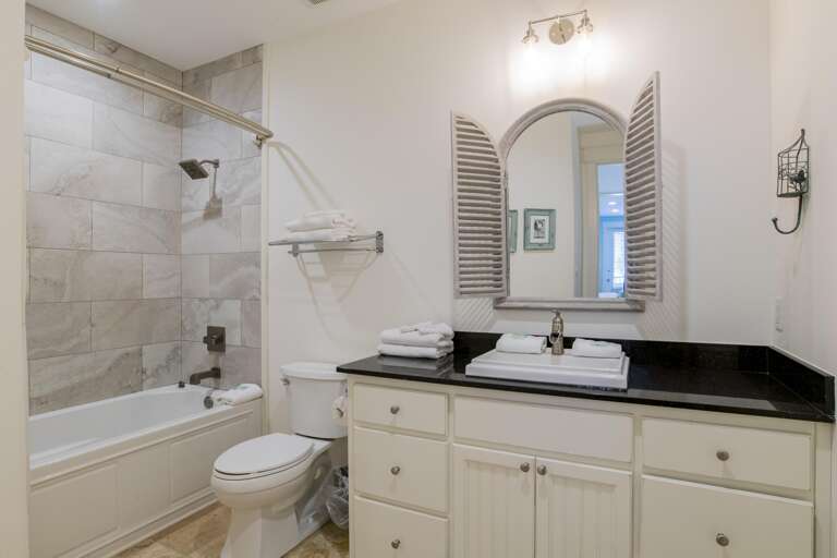 Bathtub with beige stone tile surround and oil-rubbed bronze fixtures sits beside white toilet and black granite vanity.
