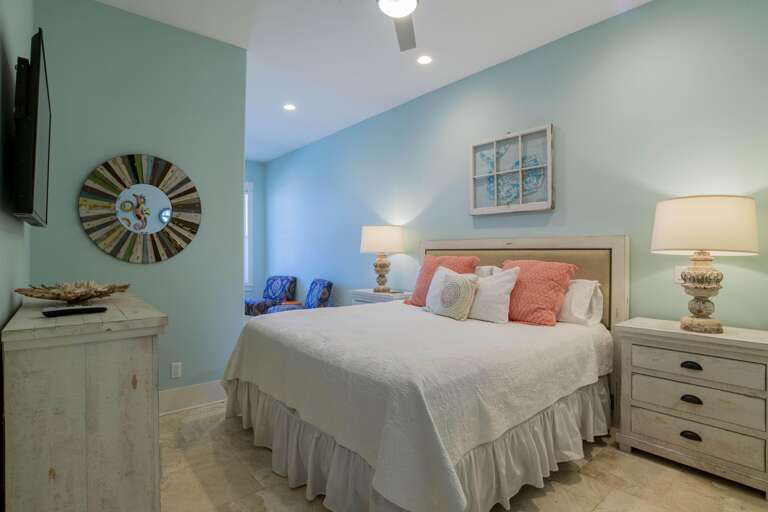 Light blue walls frame a king-size bed with coral and white pillows, flanked by distressed white nightstands with lamps.