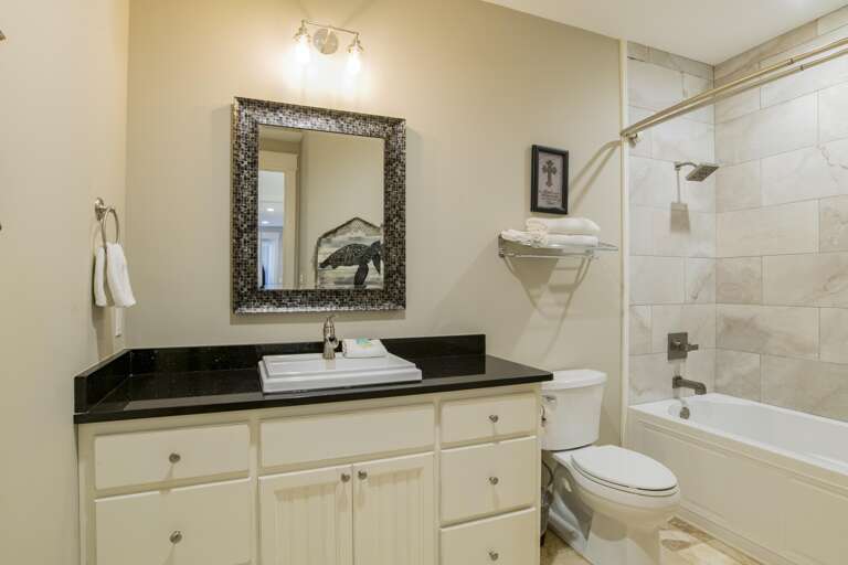 Bathroom with black granite countertop, white sink, mosaic-framed mirror, and tiled bathtub with rainfall showerhead.