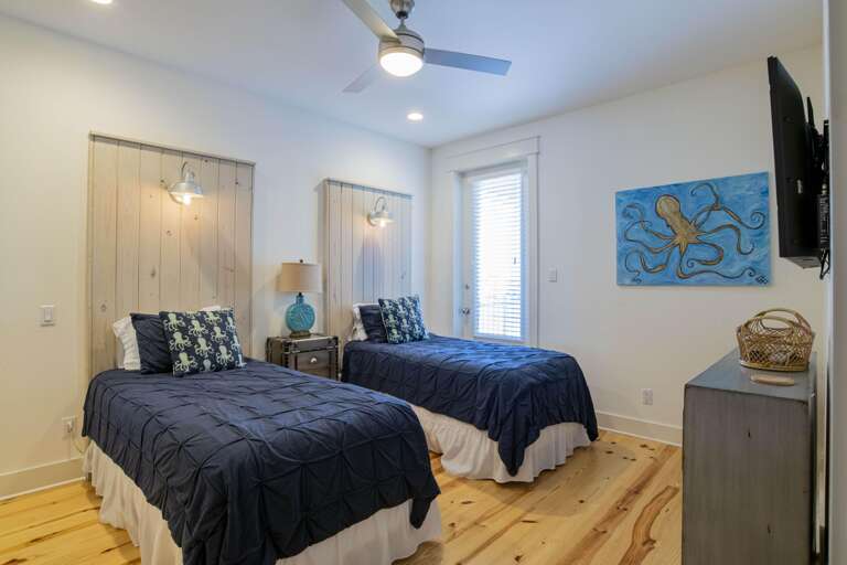 Two twin beds with navy quilts face a wall-mounted TV, accented by beach-themed pillows and artwork.