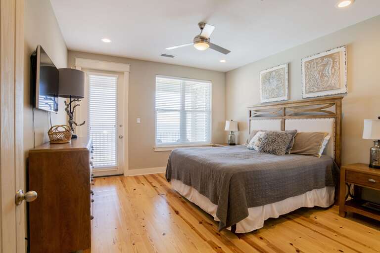 King bedroom with wooden headboard, gray quilt, polished pine floors, dresser, and door leading to balcony.