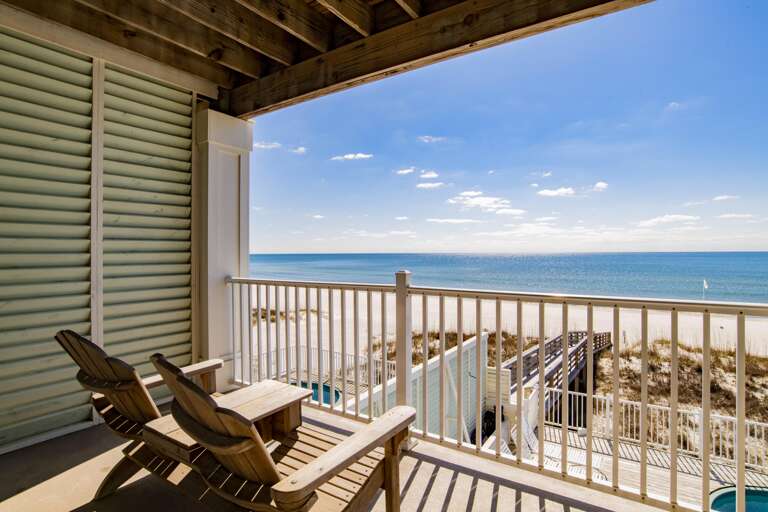Two wooden Adirondack chairs frame a sandy beach and calm Gulf waters from a shaded balcony.
