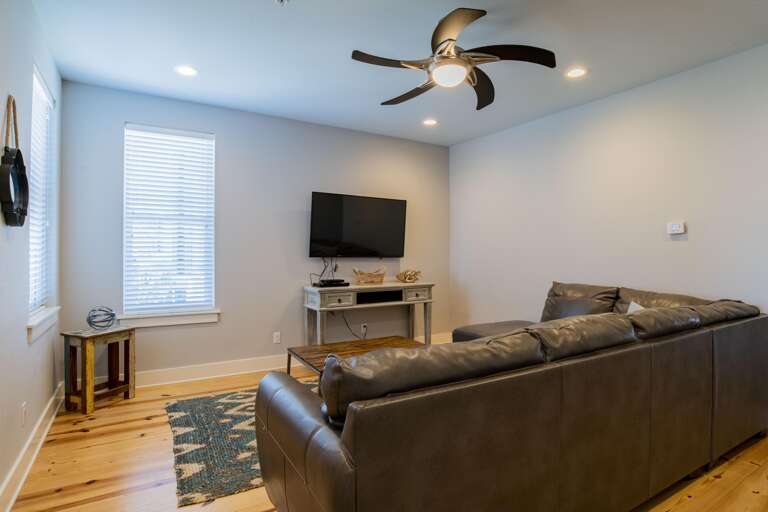 Natural light filters through blinds onto hardwood floors and a gray area rug beneath a dark leather sectional.