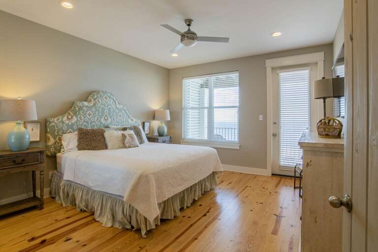 Pine floors extend beneath a window and glass door leading to a balcony, flanked by bedside tables with blue lamps.