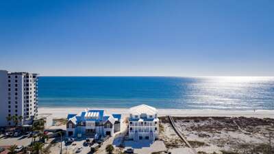 Twin beachfront homes with white roofs and wraparound balconies sit beside a sandy beach and shimmering ocean.