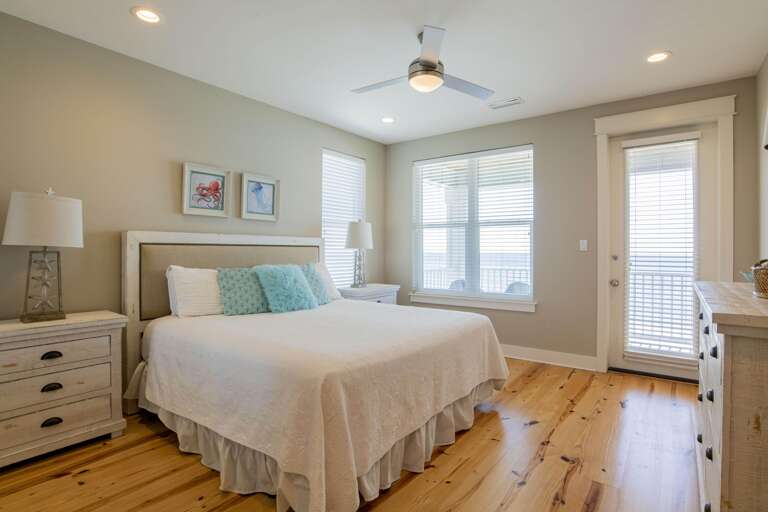 King bedroom with white quilt, soft blue pillows, pine floors, coastal lamps, and balcony access through glass door.