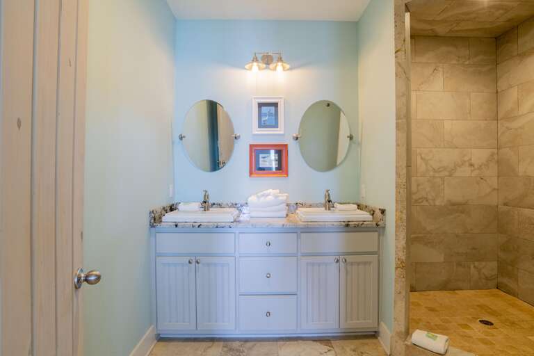Dual oval mirrors hang above a granite countertop with two white sinks in a bathroom with beige tiled walk-in shower.