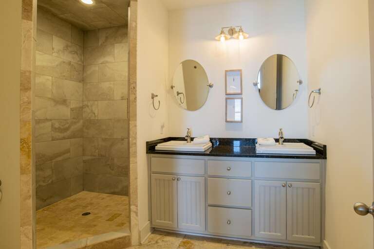 Walk-in shower with large beige stone tiles and adjacent vanity featuring brushed nickel faucets and oval mirrors.