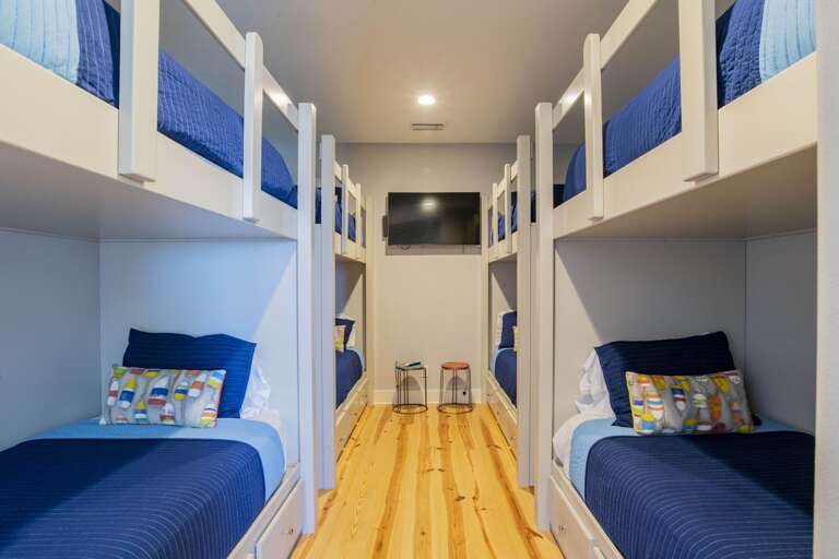 Four sets of white wooden bunk beds with blue bedding line the polished pine floor hallway beneath a wall-mounted TV.