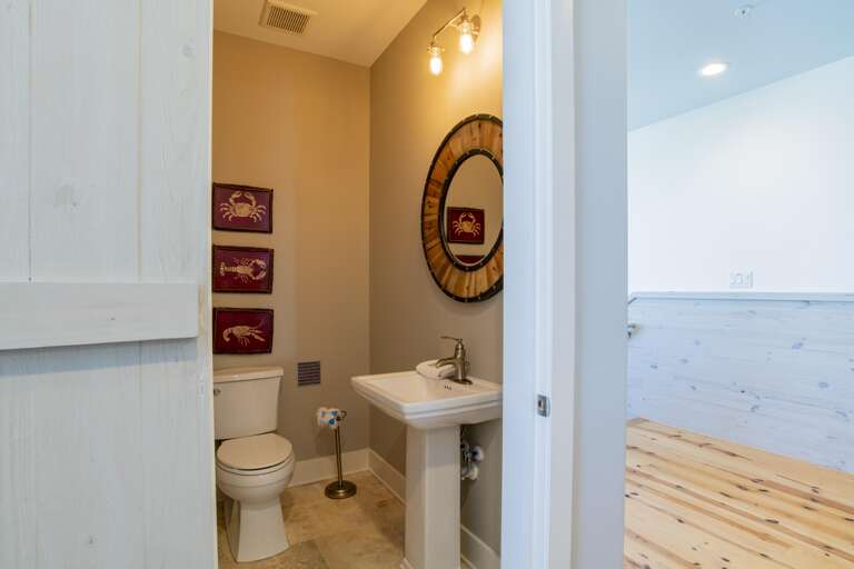 Half-open barn door reveals a small bathroom with beige walls, pedestal sink, round wood-framed mirror, and coastal artwork.