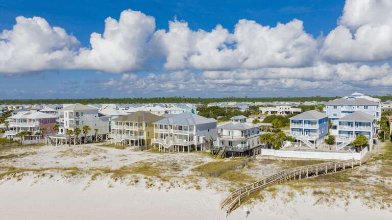 Beachfront homes with raised wooden decks and boardwalks lead across white sand dunes under a blue sky.