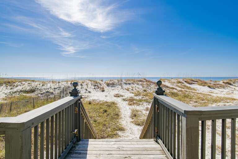 Wooden staircase with dark railings leads down through sand dunes to a bright blue Gulf shoreline under a wispy cloud sky.