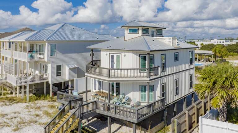 Beach house exterior with white siding, metal roof, wraparound balconies, and Adirondack chairs on the lower deck.