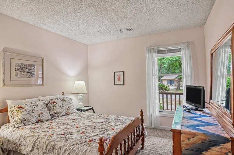 Your cozy bedroom features a comfortable bed dressed in botanical linens, soft natural light from the window, and serene views of the leafy neighborhood beyond.