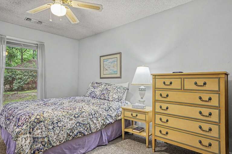 Step into your peaceful bedroom retreat with a comfy bed dressed in charming linens, classic furnishings, a gentle ceiling fan, and a window framing leafy garden views.