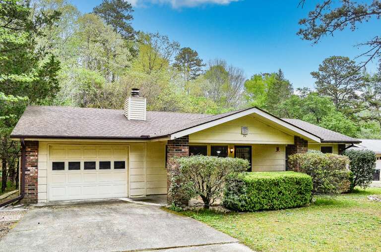 A charming single-story home nestled among mature trees and lush landscaping, featuring a spacious driveway and serene natural surroundings.
