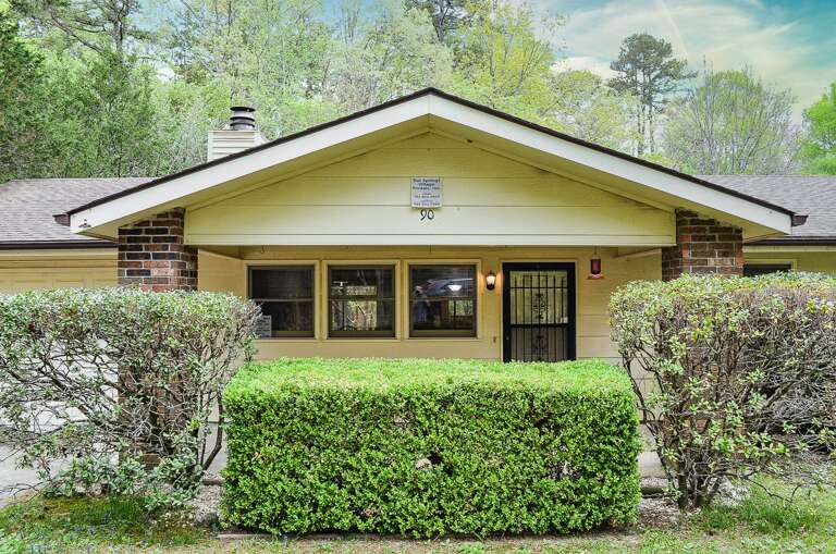 Charming yellow cottage with brick accents, nestled in lush greenery and mature trees, offering a peaceful private retreat surrounded by nature.