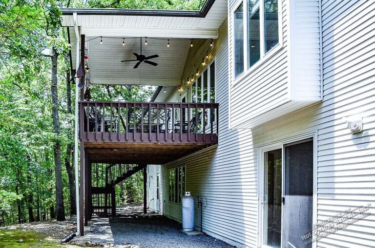 This wooded property features a spacious upper-level deck with ambient string lights and ceiling fan, perfect for enjoying the surrounding forest. Ground-level parking area and exterior entrance provide convenient access.