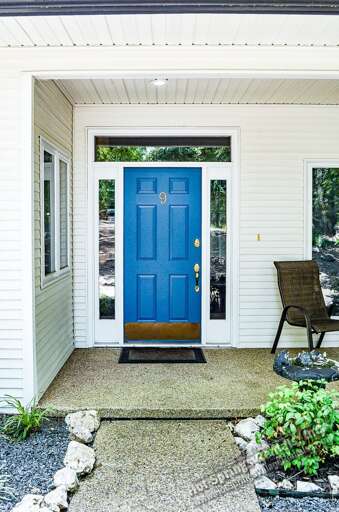 A welcoming blue front door opens to a bright, covered porch framed by white siding and natural greenery, setting the tone for a relaxed getaway.