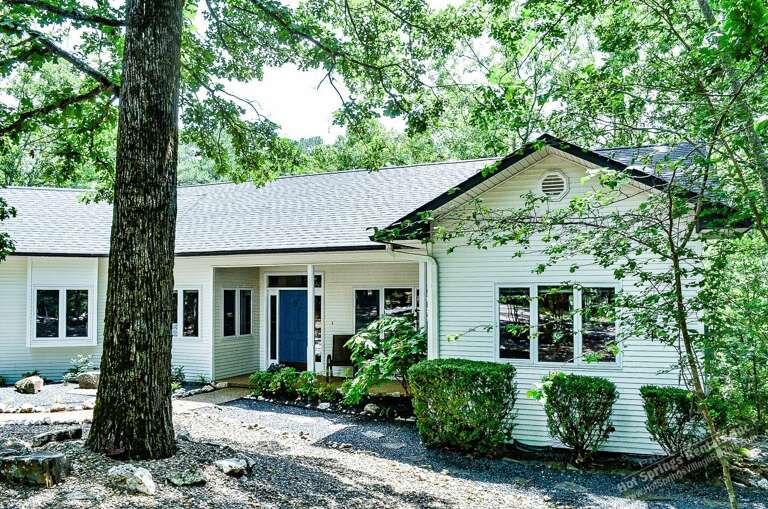 A welcoming two-story vacation home with a distinctive blue front door nestled in a peaceful wooded setting, surrounded by mature trees and natural landscaping.