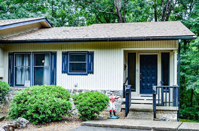 This charming woodland cottage features a welcoming front entrance surrounded by lush greenery and nestled among mature trees. The stone-accented exterior and natural setting create a peaceful retreat atmosphere perfect for your getaway.