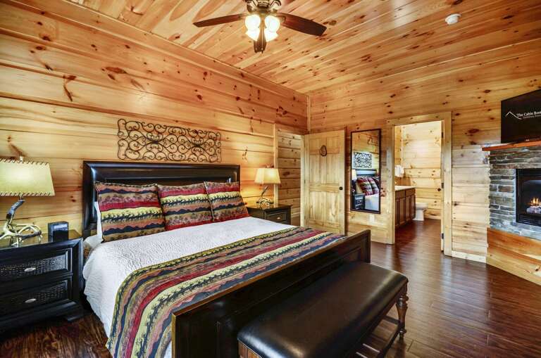 Bedroom with a large bed, bench at foot of the bed, and a pair of nightstands, all in front of a fireplace.