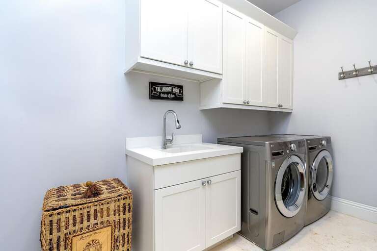 A dedicated laundry room includes a full-size washer and dryer, utility sink, countertop, and white storage cabinets.
