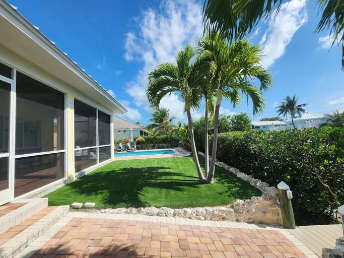 A manicured turf lawn, mature palm trees, tropical hedges, and brick pathways surround the outdoor living area.