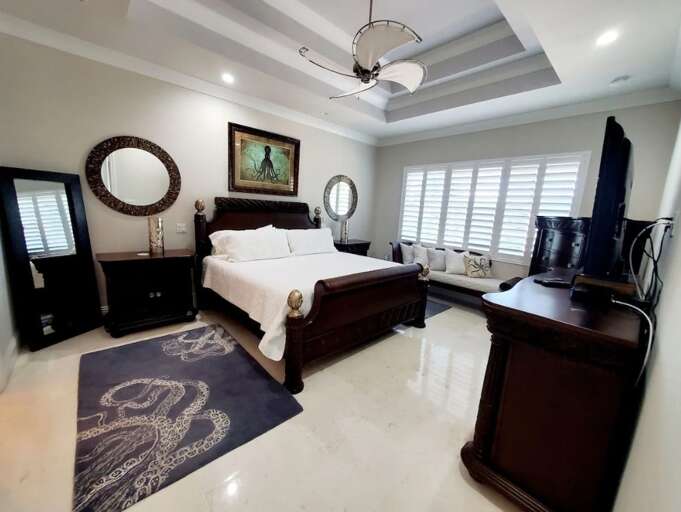 The primary bedroom offers a comfortable bed, dark wood furnishings, a tray ceiling, television, and abundant natural light.