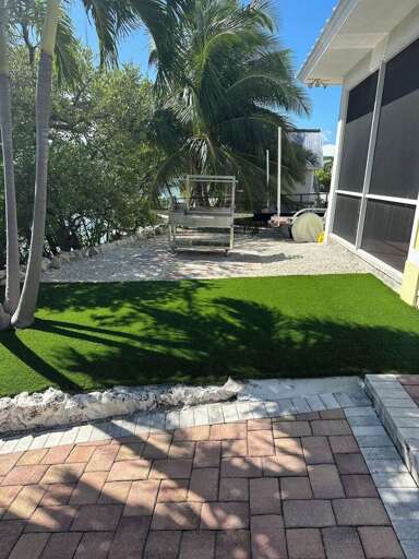 This shaded side yard features low-maintenance artificial turf, tropical surroundings, brick pathways, and a wood-burning grill