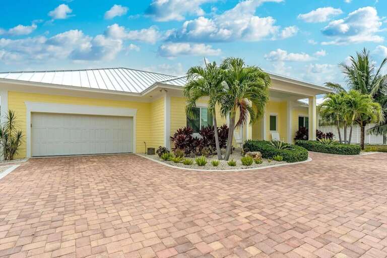 The sunny yellow home features an attached garage, expansive brick driveway, palm trees, and manicured tropical landscaping.