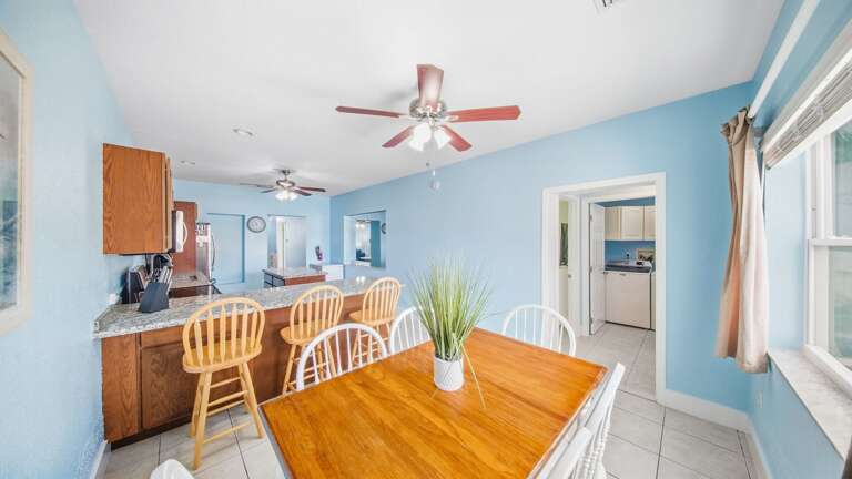 This alternate view showcases the wooden dining table, kitchen breakfast bar, tiled floors, and cheerful coastal-blue walls.