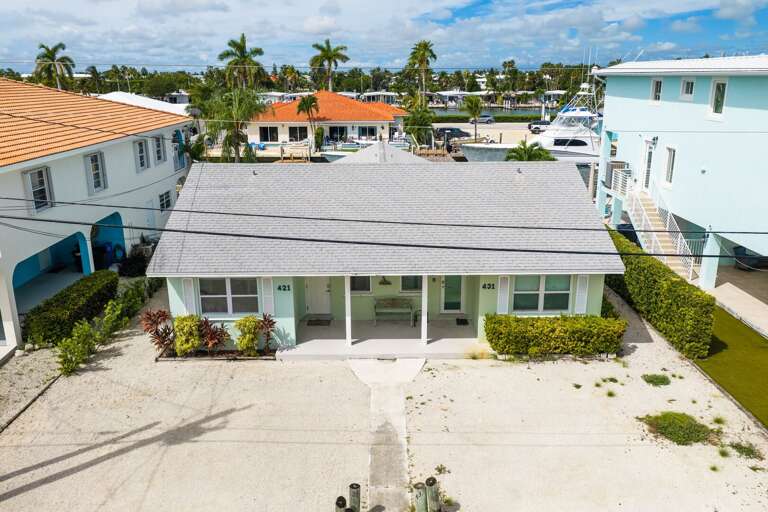 An elevated front view highlights the ground-level home, covered entrance, tropical landscaping, and spacious parking area.