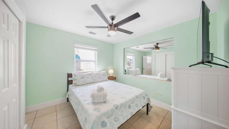 This inviting bedroom includes a comfortable bed, large wall mirror, white furnishings, ceiling fan, and abundant natural light.