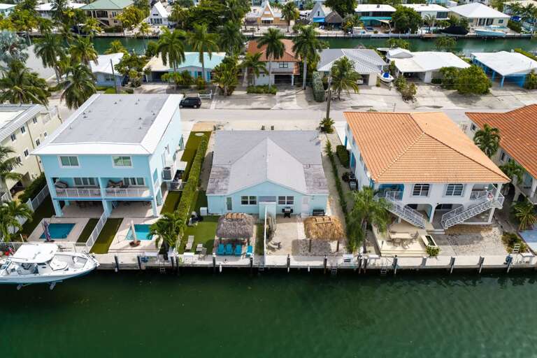 This waterfront aerial view highlights the private dock, tropical backyard, tiki areas, and direct boating access.