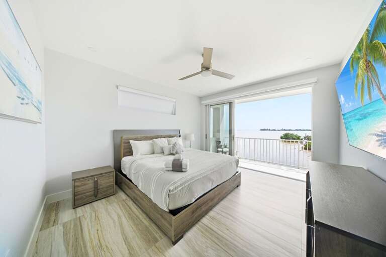 This inviting primary bedroom features a cozy King bed, coastal furnishings, a ceiling fan, and sliding glass doors opening to a private waterfront balcony.