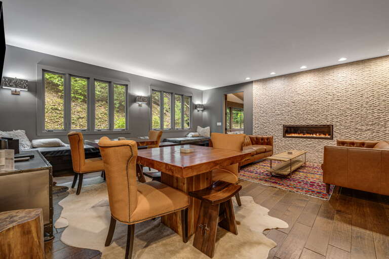 Gather around the rich wood table in your spacious living area, where natural light streams through tall windows revealing lush greenery outside. Sink into plush leather seating by the modern fireplace and feel completely at home.