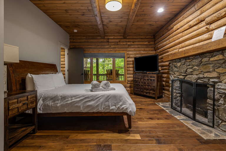 Unwind in your cozy log cabin bedroom, where rustic timber beams and a stone fireplace create a warm mountain retreat. The glass doors open to lush forest views, and crisp linens await your comfort.